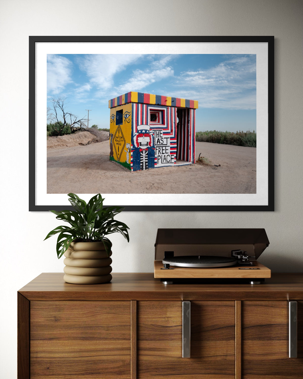 Slab City “The Last Free Place” photo print above a record player and plant on a wooden cabinet.