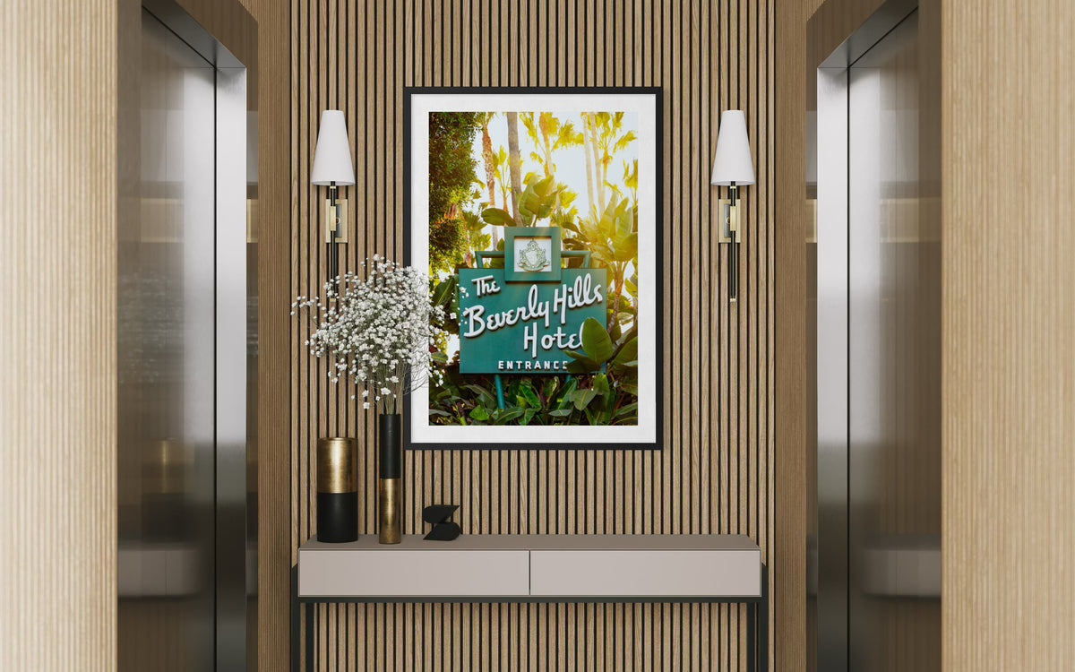 Beverly Hills Hotel sign in a frame mounted between elevators, above a console with vases and flowers.