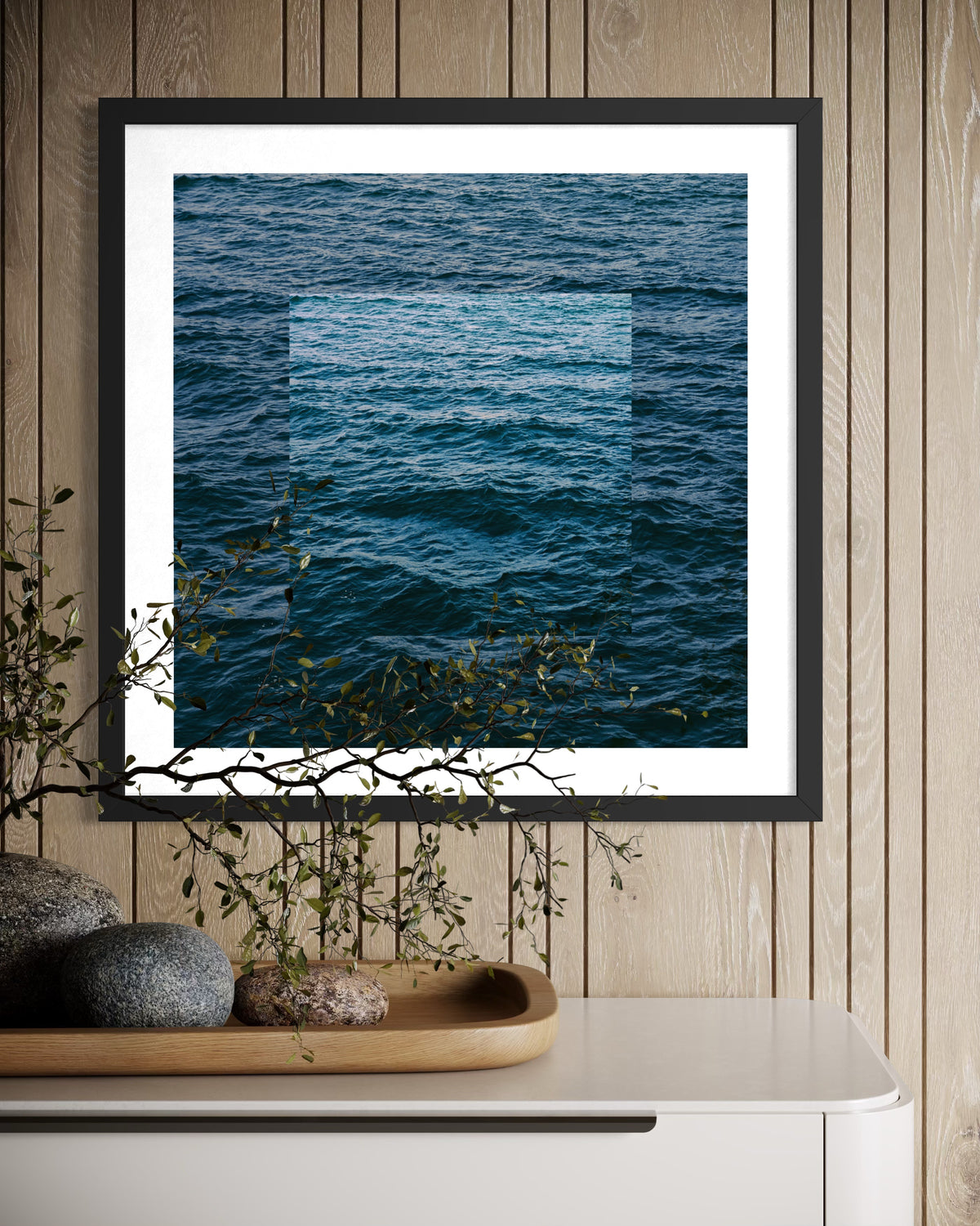 Framed abstract blue ocean print above a white table with stones and a leafy branch tray, set against wooden walls.
