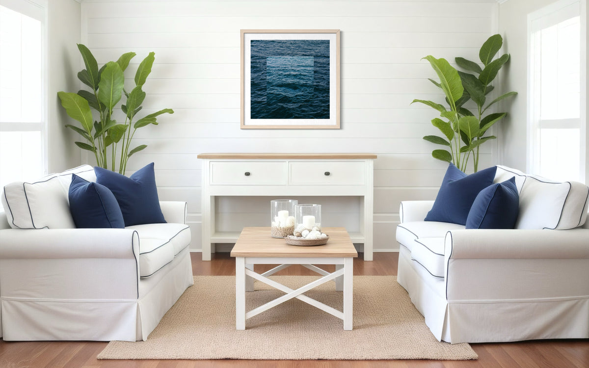 White sofas with navy pillows, indoor plants, and Blue Ocean wall art hanging above the seating area in a living room.