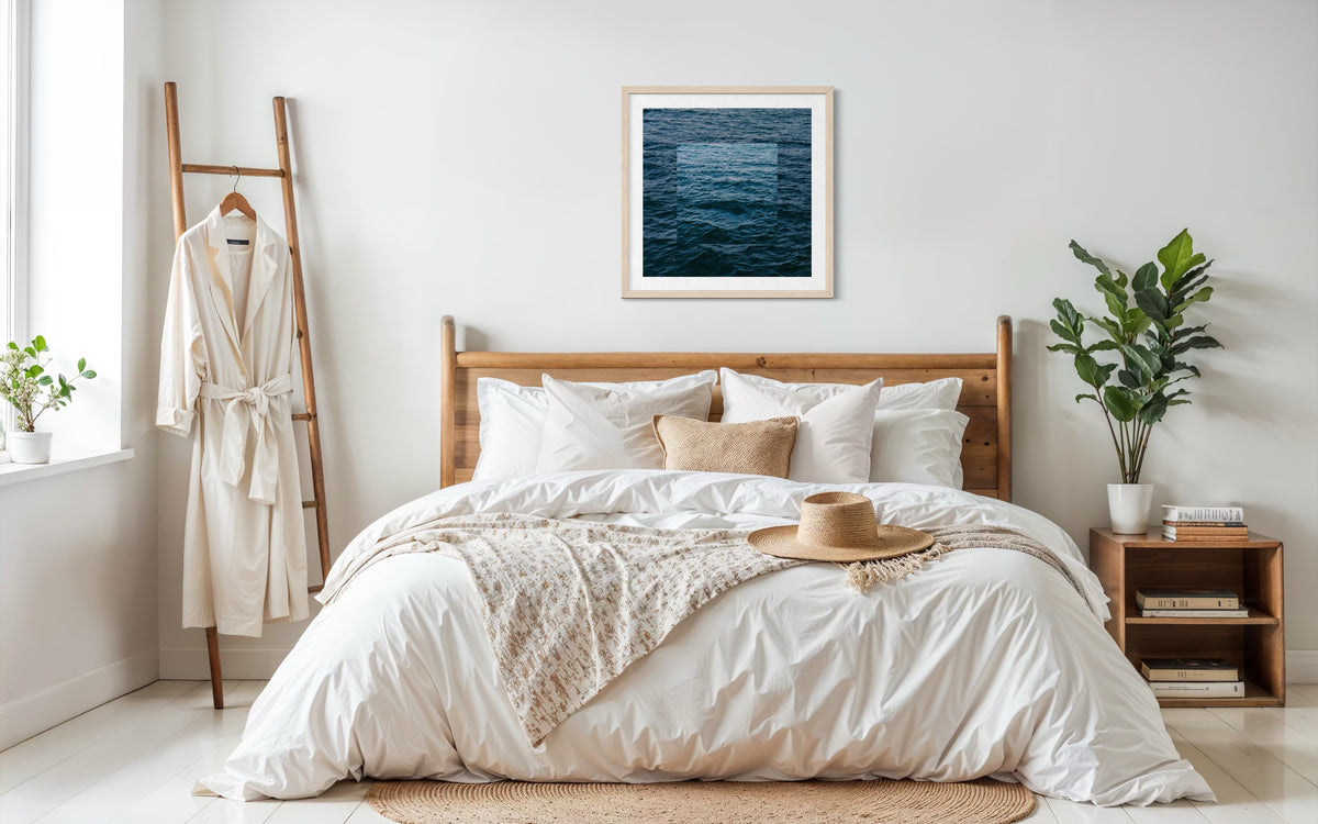 Wooden bed with white bedding, robe, plants, and Blue Ocean wall art in a cozy bedroom setting.