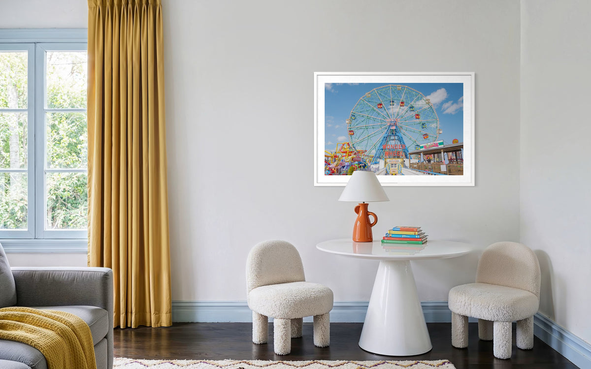 Coney Island Wonder Wheel photograph on a light gray wall above a round table with white chairs and a lamp.