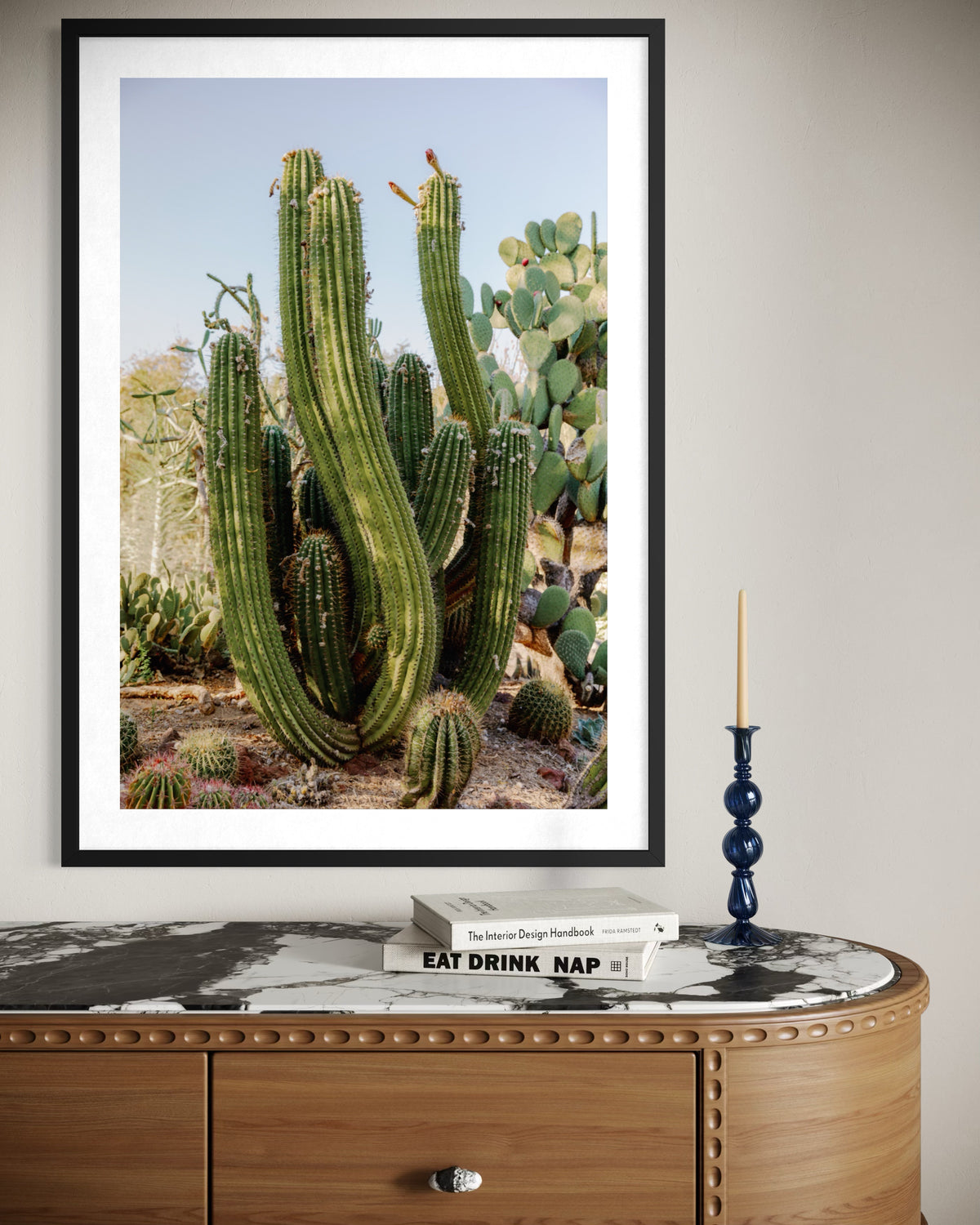 Framed print of an elephant cactus above a marble console table with books, succulents, and a blue candle holder.