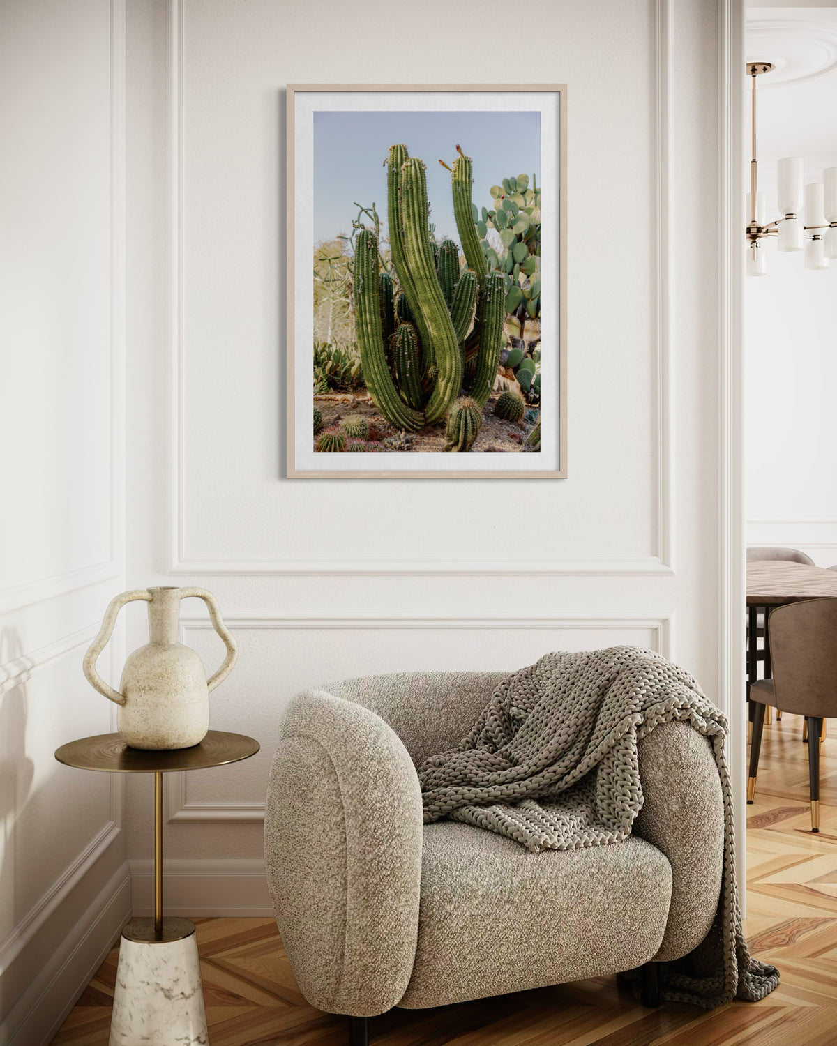 Beige armchair with gray blanket beside small table and vase, under framed Elephant Cactus photo on white wall.