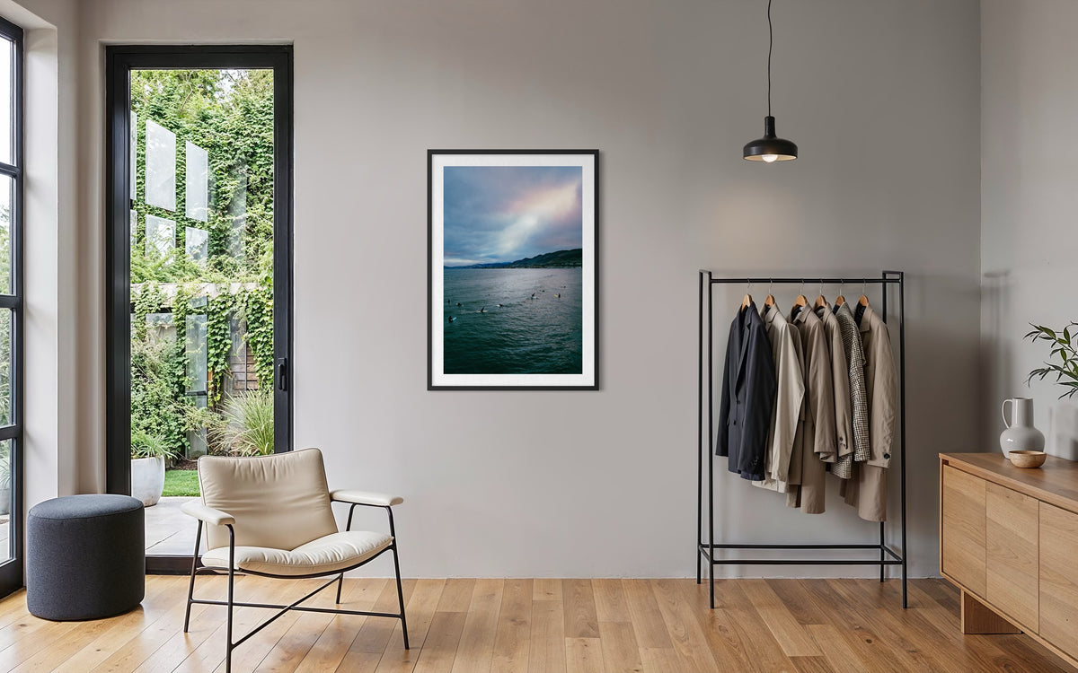 Framed “Ethereal Sky, Pismo Beach” photo on wall above chair, large window, and clothes rack in modern room.