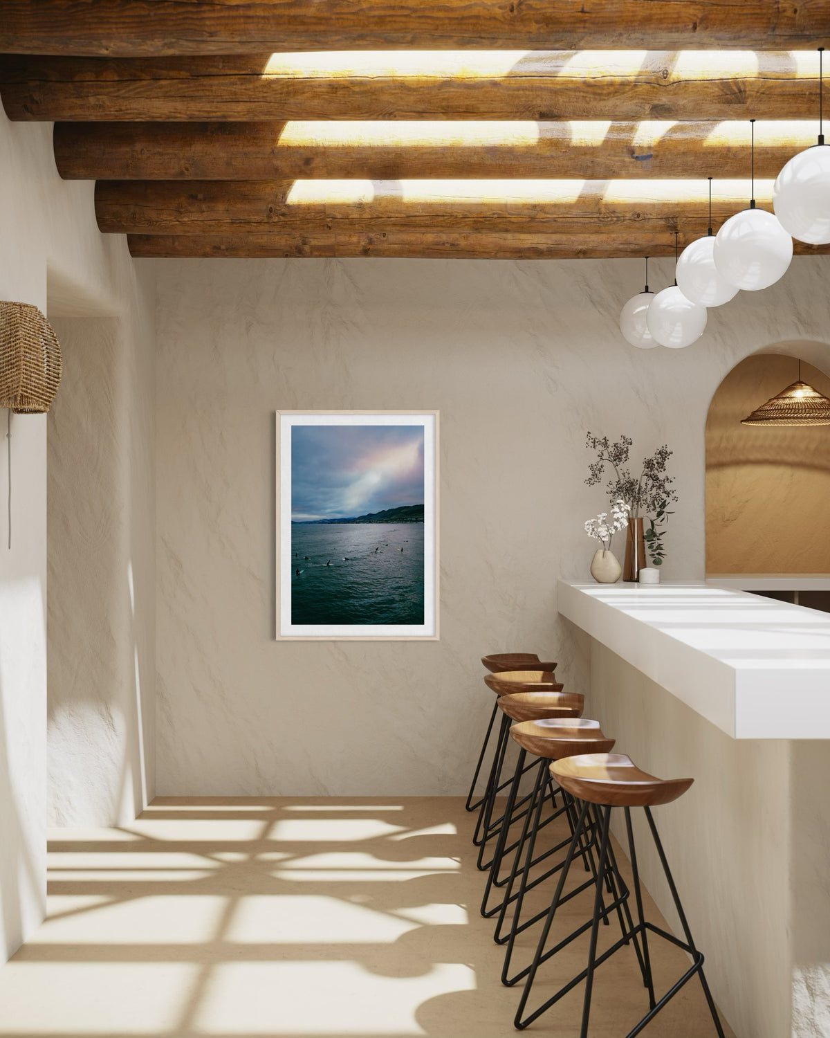 Modern kitchen with wood beams, globe lights, bar stools, and “Ethereal Sky, Pismo Beach” art on beige wall.