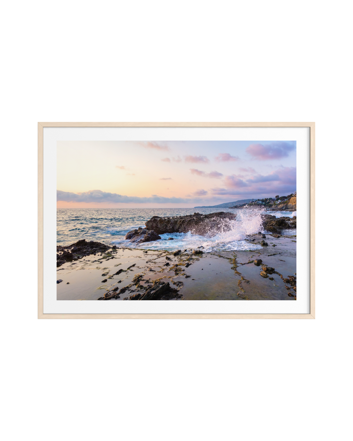 Waves crash on Laguna Beach’s rocky shore at sunset, with pastel skies and a light wood frame visible.