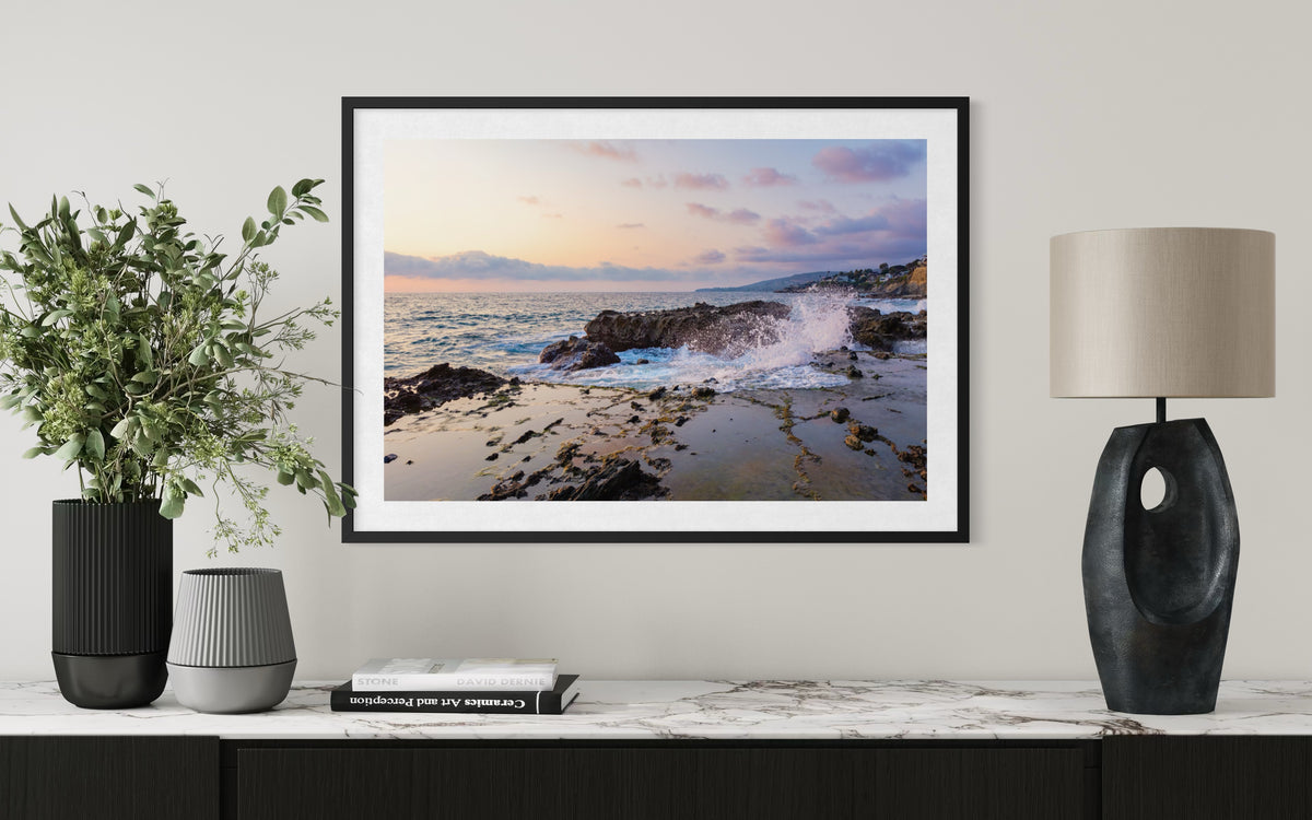 Framed ocean photograph titled “Evening Splash” displayed above a marble console, with plants and a lamp nearby.