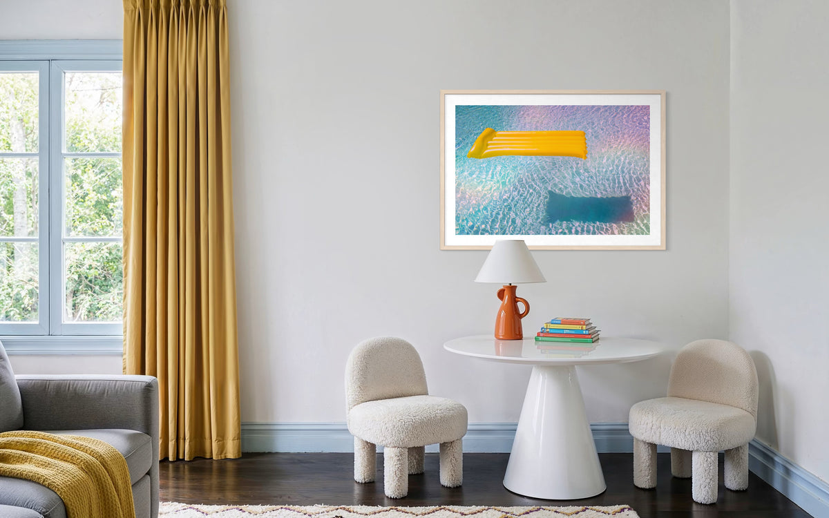 Round table and two chairs beside lamp; books under framed “Floating Through Summer” yellow pool float print.
