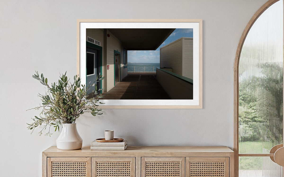 Framed Harbor Office ocean view photograph above cabinet with vase, books, and arched window in natural light.