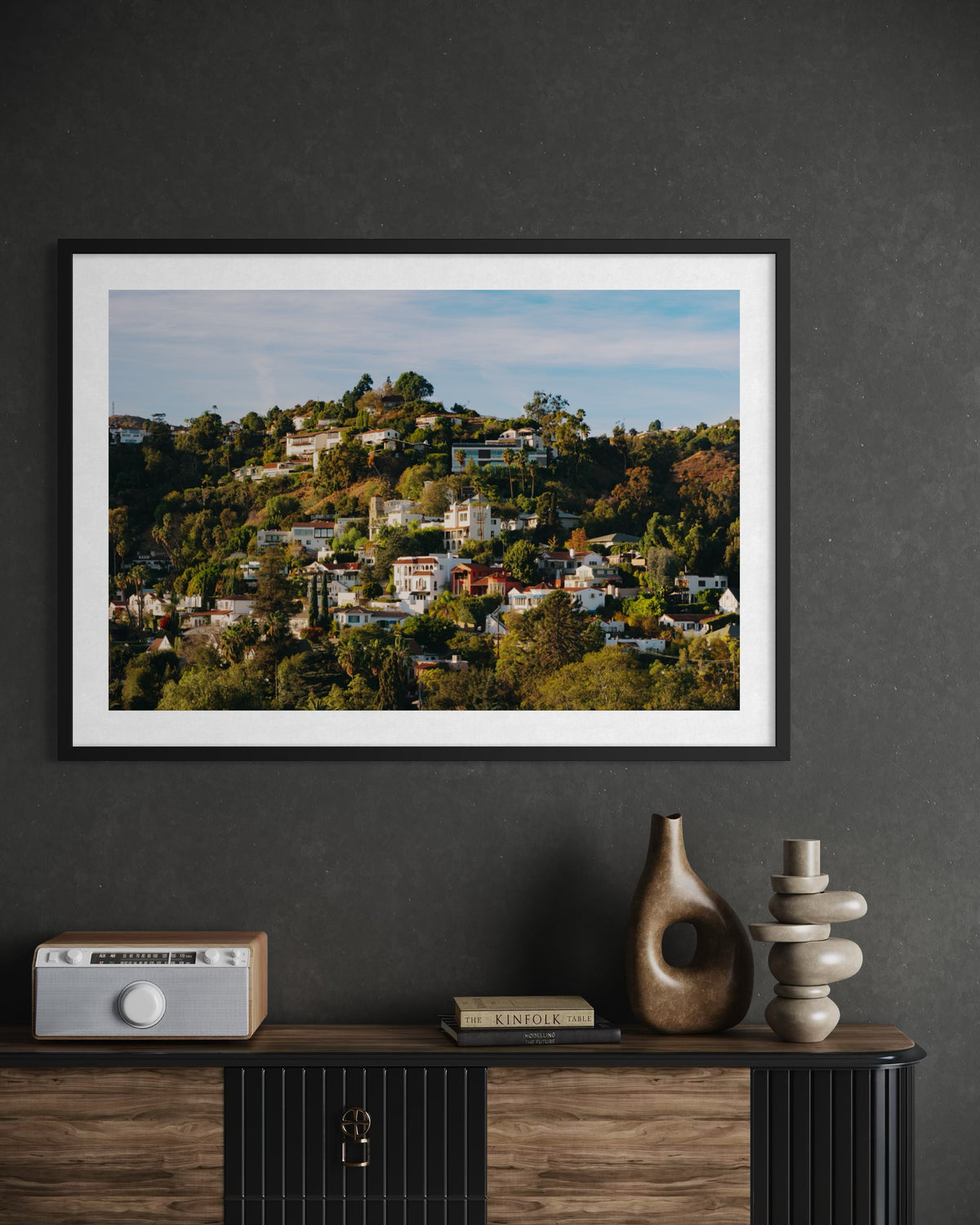 Framed “Hollywood Hills” Los Angeles photo above a dark cabinet holding vases, books, and a radio.