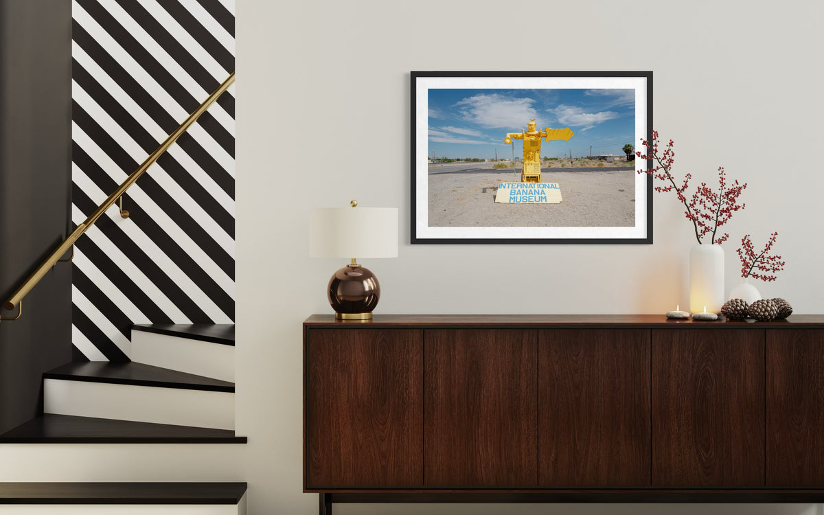 Framed “International Banana Museum” sign photograph hangs above wooden console, desert-themed decor in view.