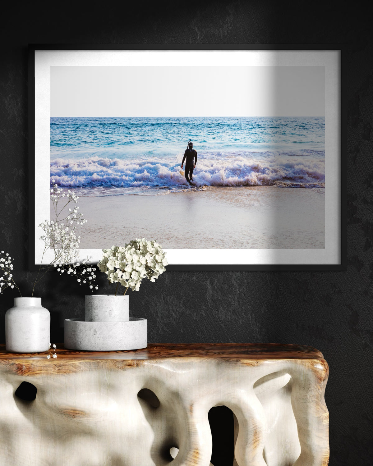 Framed print of a lone person by the Pacific Ocean, titled “Into The Sea,” displayed above a table with white vases.