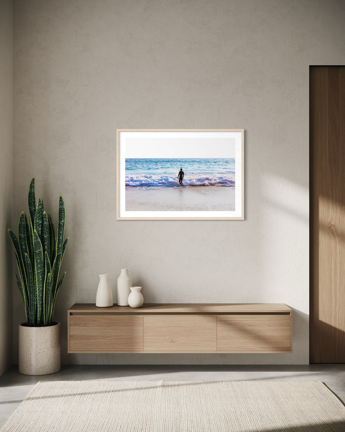Framed beach print titled “Into The Sea” with a lone figure, displayed above plant and vases in a simple room.