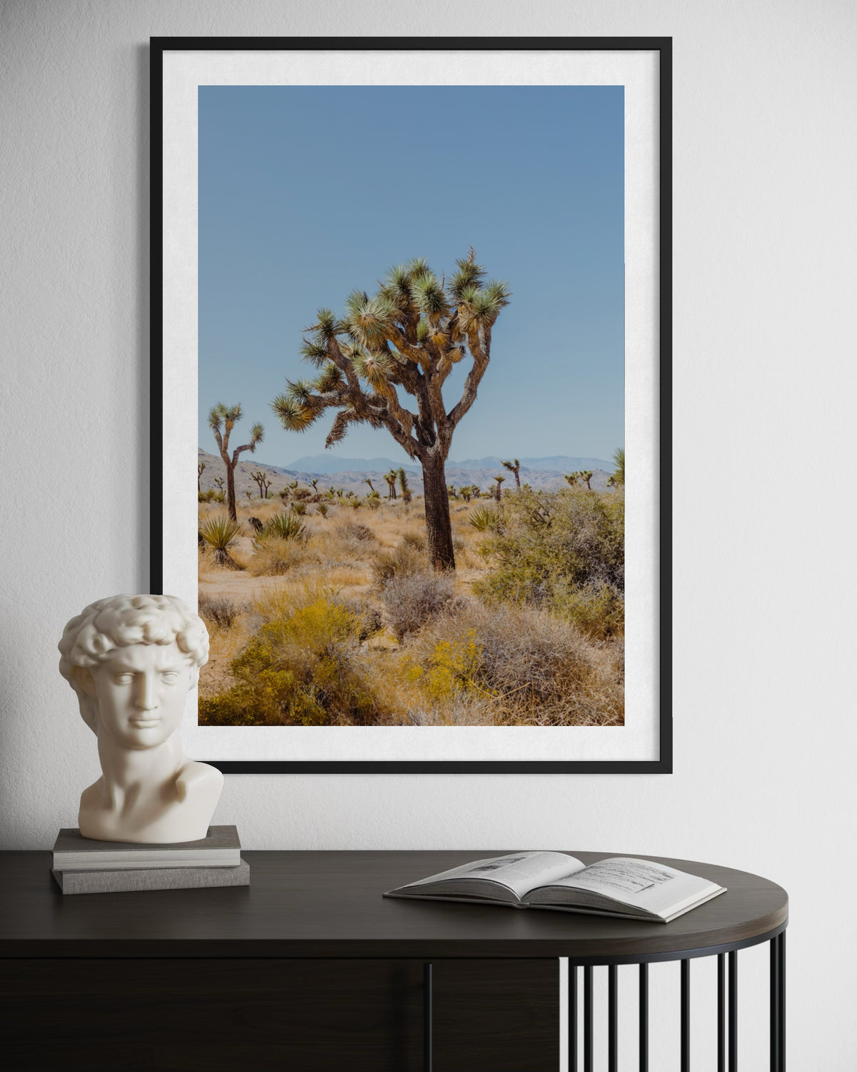 Framed print of Joshua Tree desert landscape displayed above a bust and open book on a dark tabletop.