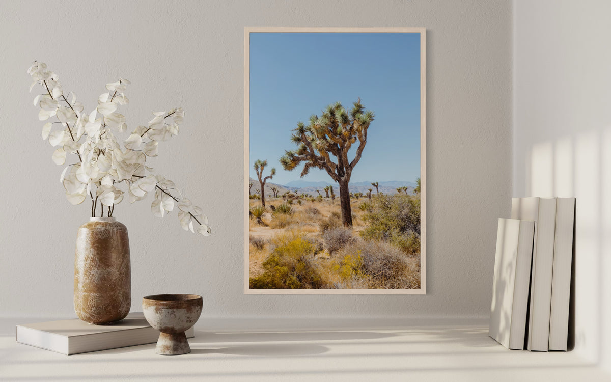 Framed Joshua Tree II desert landscape print on light table with books and white flower vases arranged nearby.