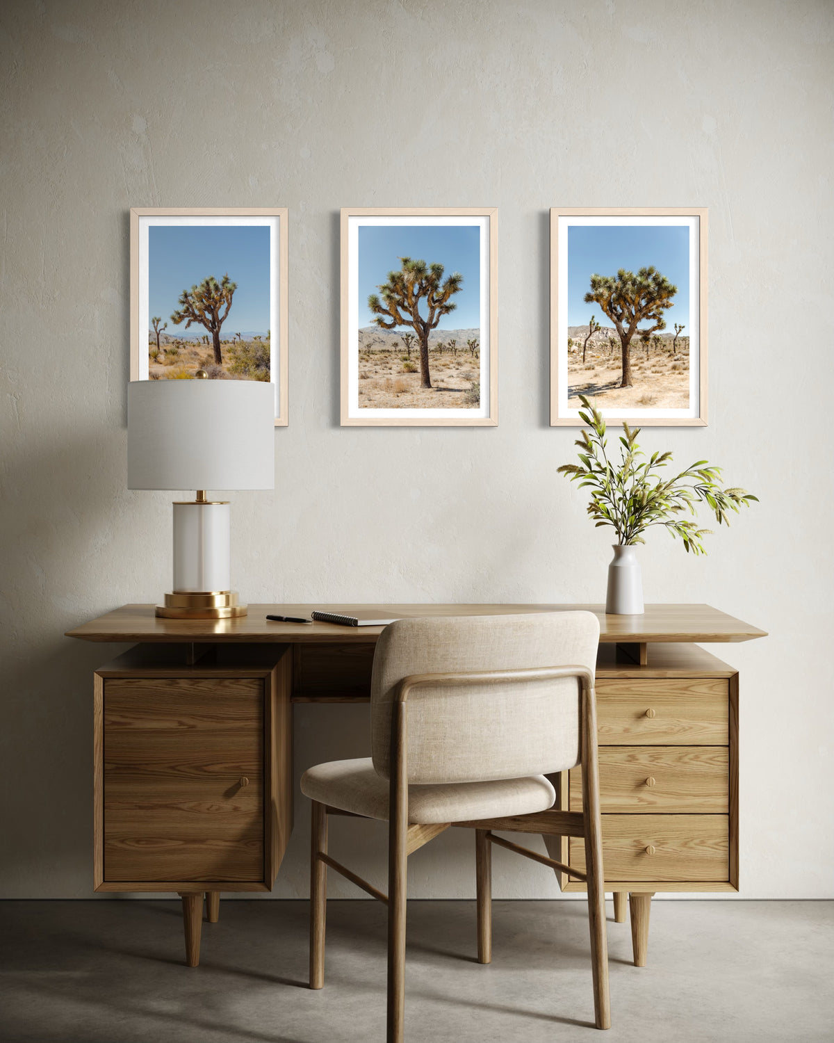 Framed Joshua Tree photographs on beige wall above a wooden desk with chair, white lamp, and green plant.