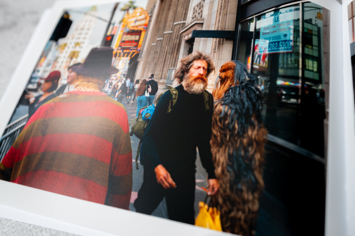 A bearded man with a backpack walks by people in costumes on an LA street.