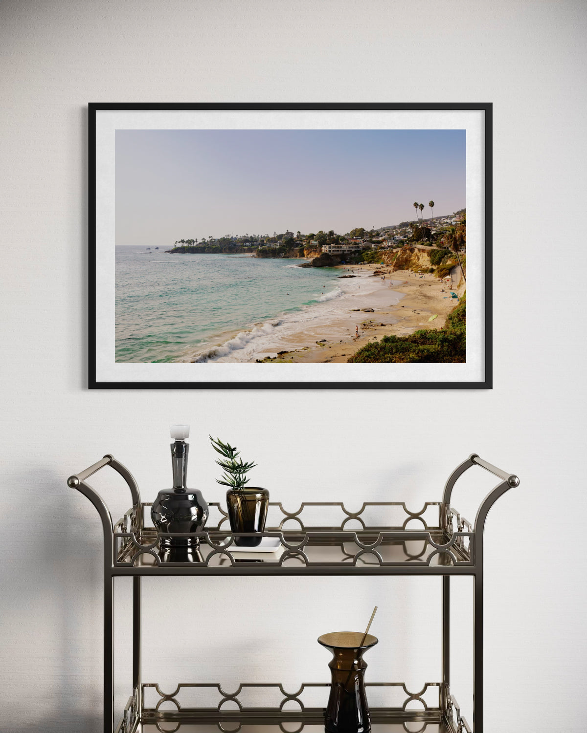 Framed Laguna Beach, California print above a bar cart with bottles, glasses, and a plant against a white wall.