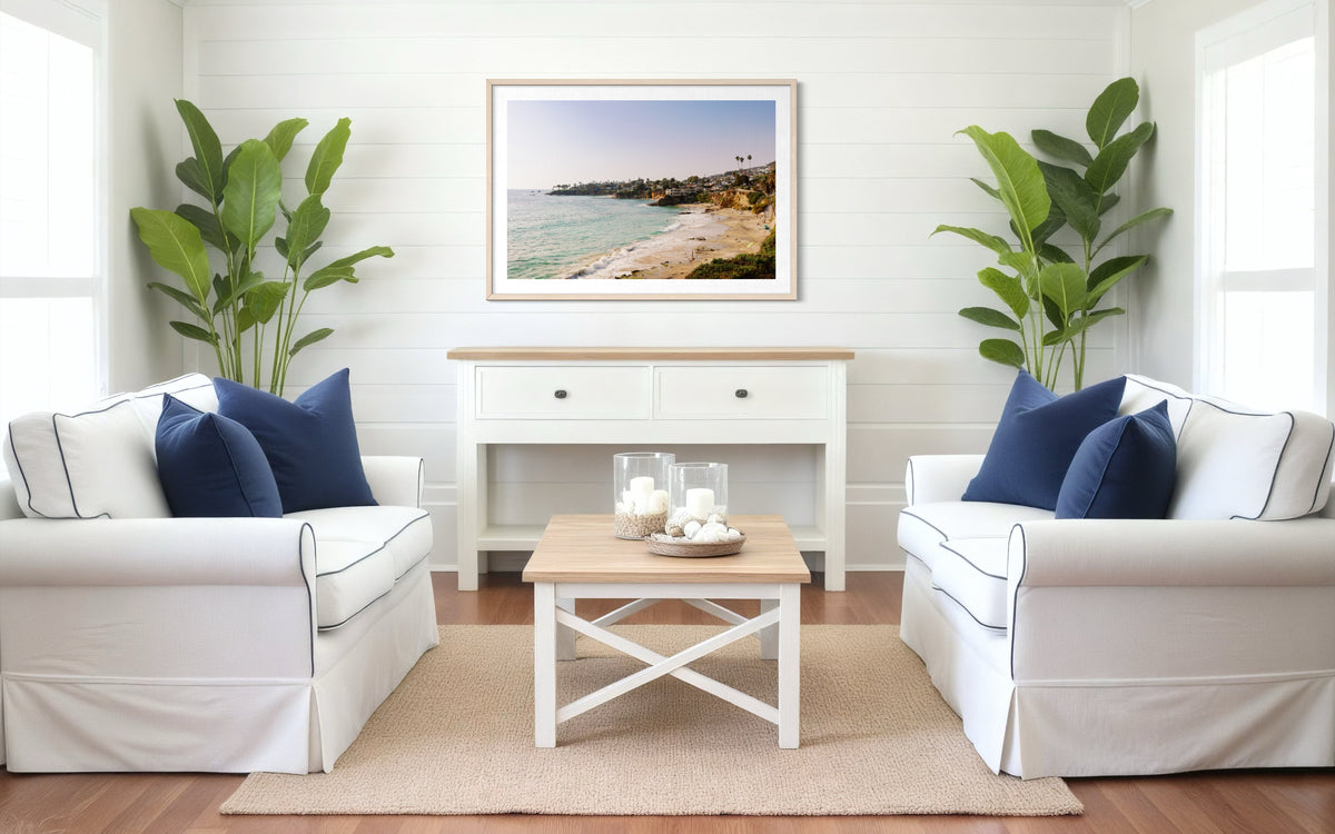 Laguna Beach View artwork hangs above a console in a bright living room with white furniture, blue pillows, and plants.