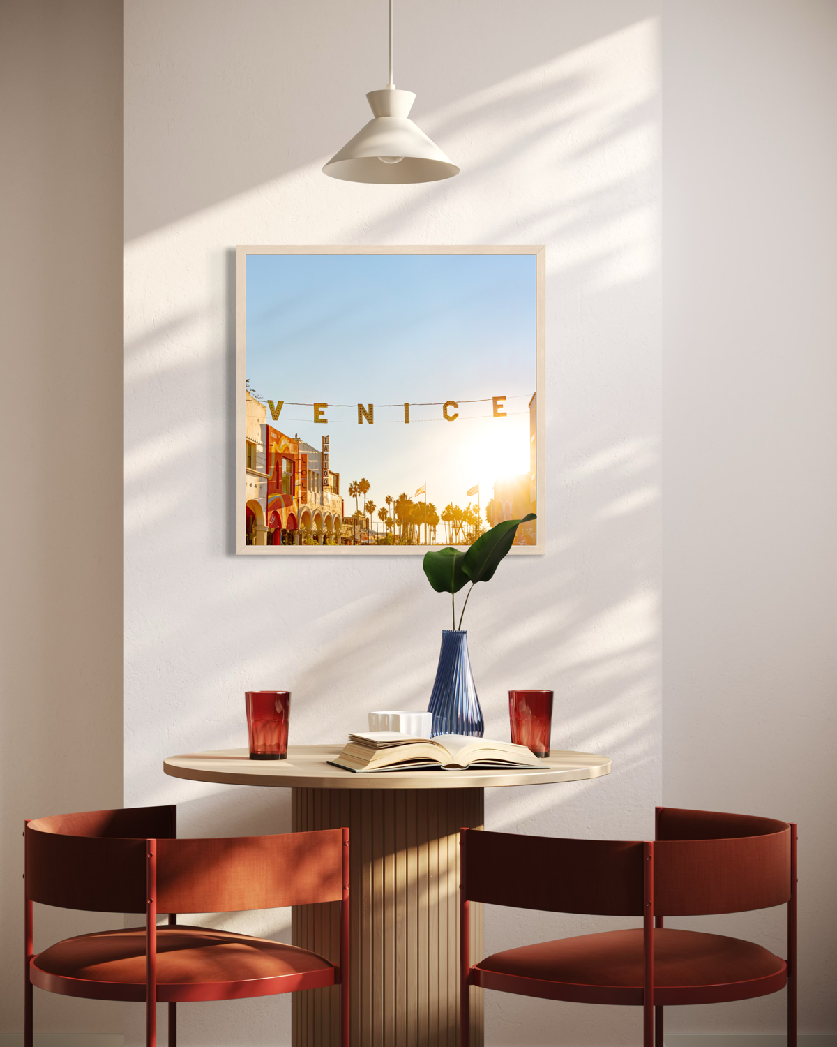 Two red chairs and a table with two red glasses beneath “Magic Hour in Venice” artwork, set indoors.