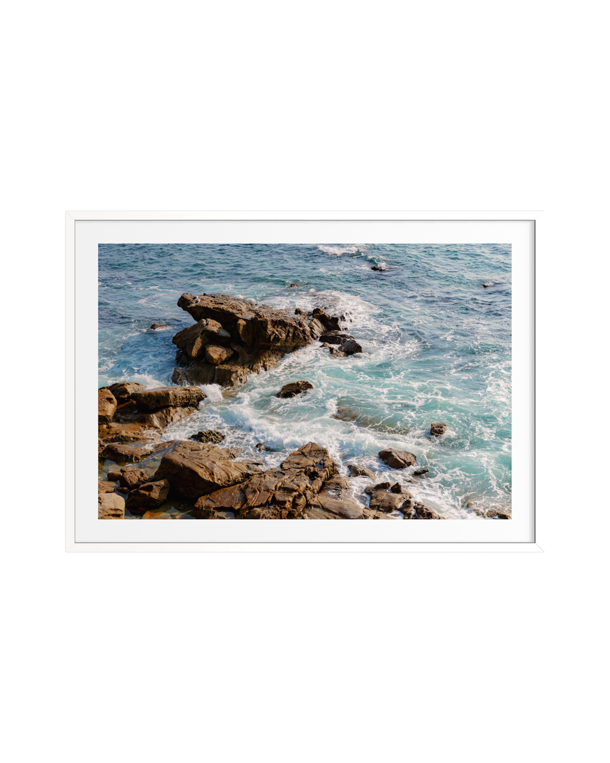 Turquoise waves crash against brown rocks on a rocky shoreline in bright daylight.