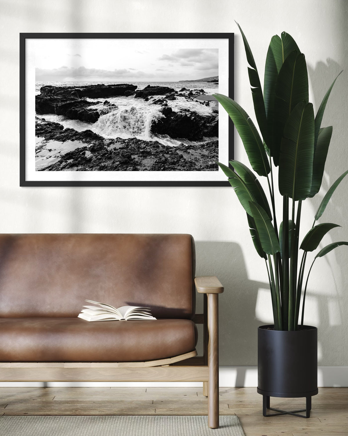 Brown leather sofa, open book, and potted plant beneath “Ocean Momentum” ocean photo on a white wall.
