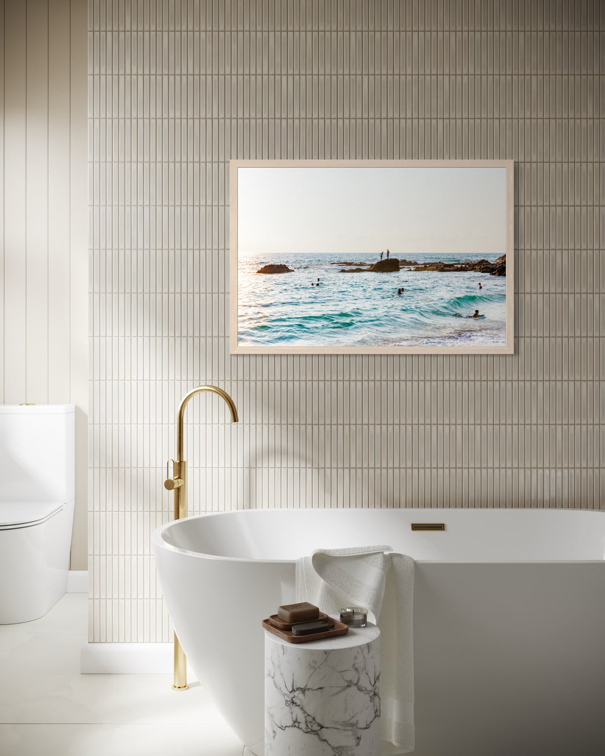 White freestanding tub, gold faucet, marble table, and “Ocean Swim” photo on beige tile bathroom wall.