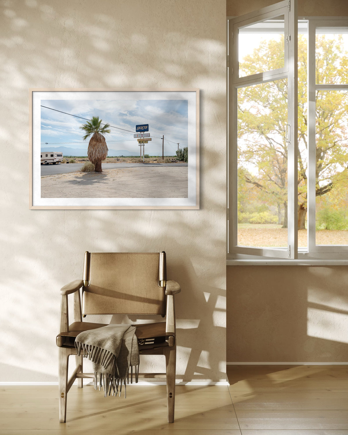 Beige chair with blanket under Palm Desert roadside art, sunny window, and autumn trees outside.