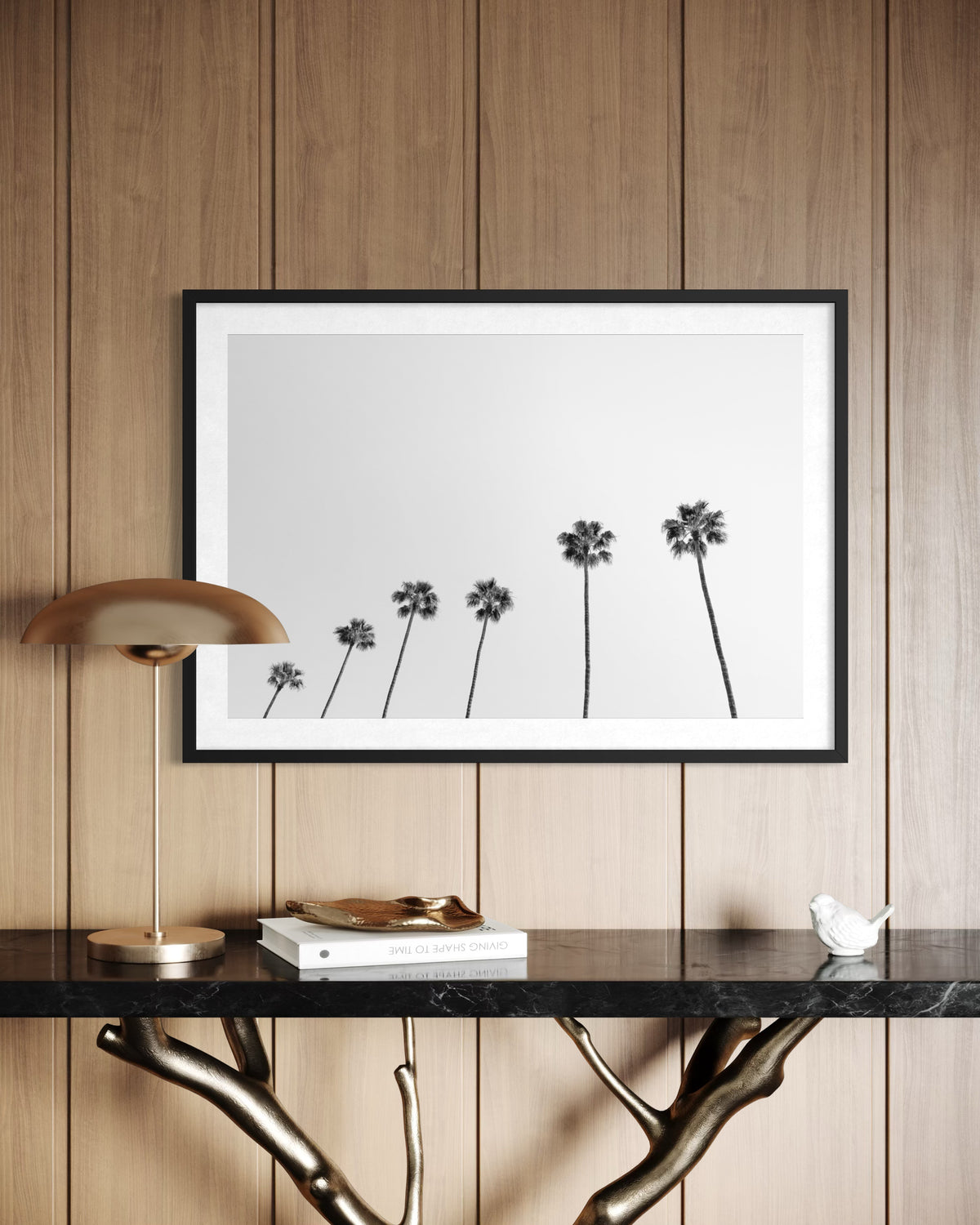 Framed print of palm trees hangs above a black table and lamp, set against wood-paneled walls.