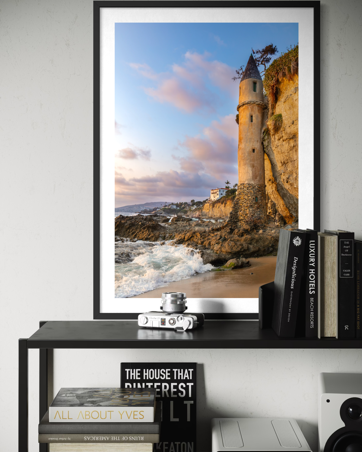 Framed print of Pirate Tower in Laguna Beach displayed on a shelf among books, magazines, and a camera.