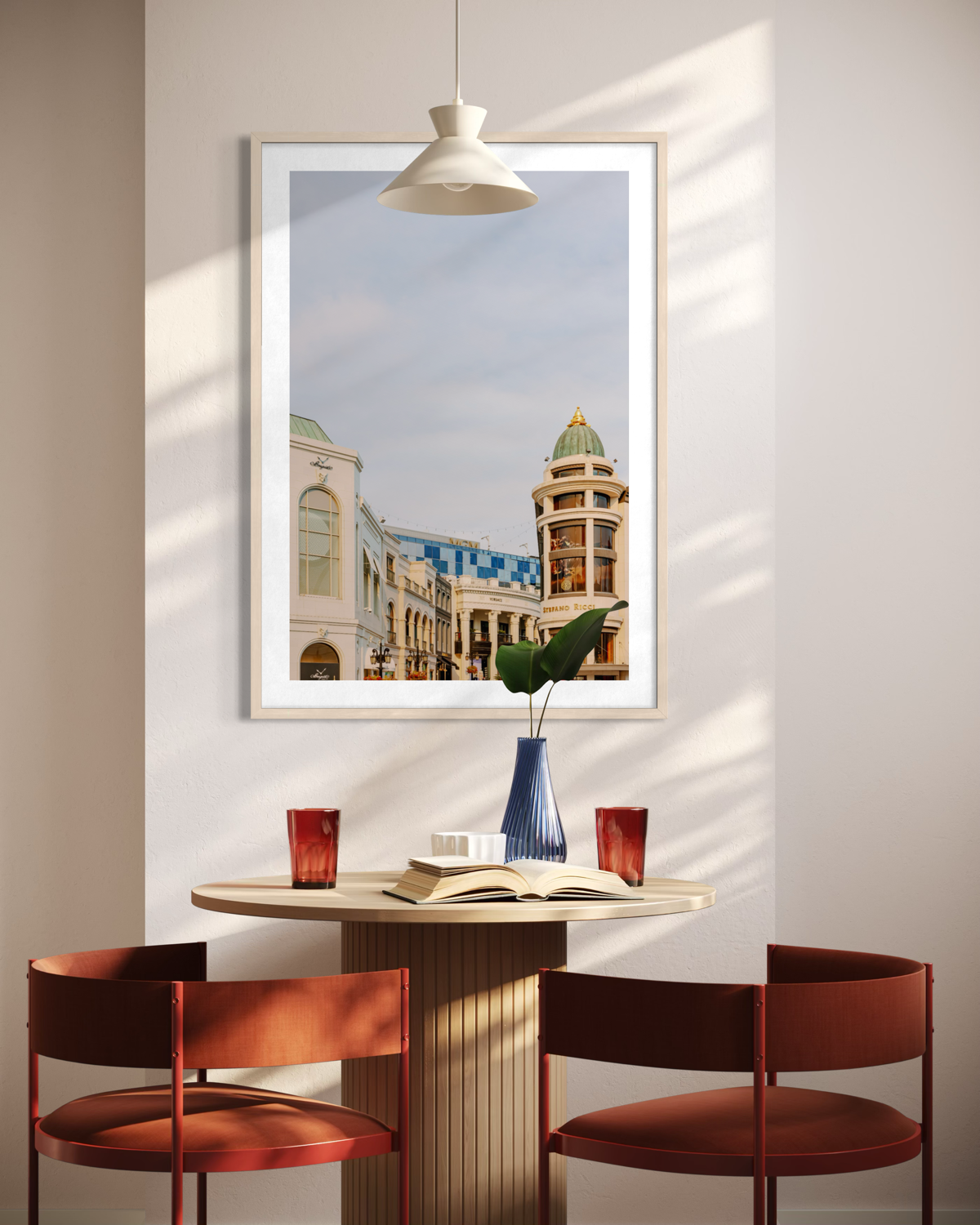 Framed Rodeo Drive cityscape hangs above a round table with two red chairs, a vase, and glasses.