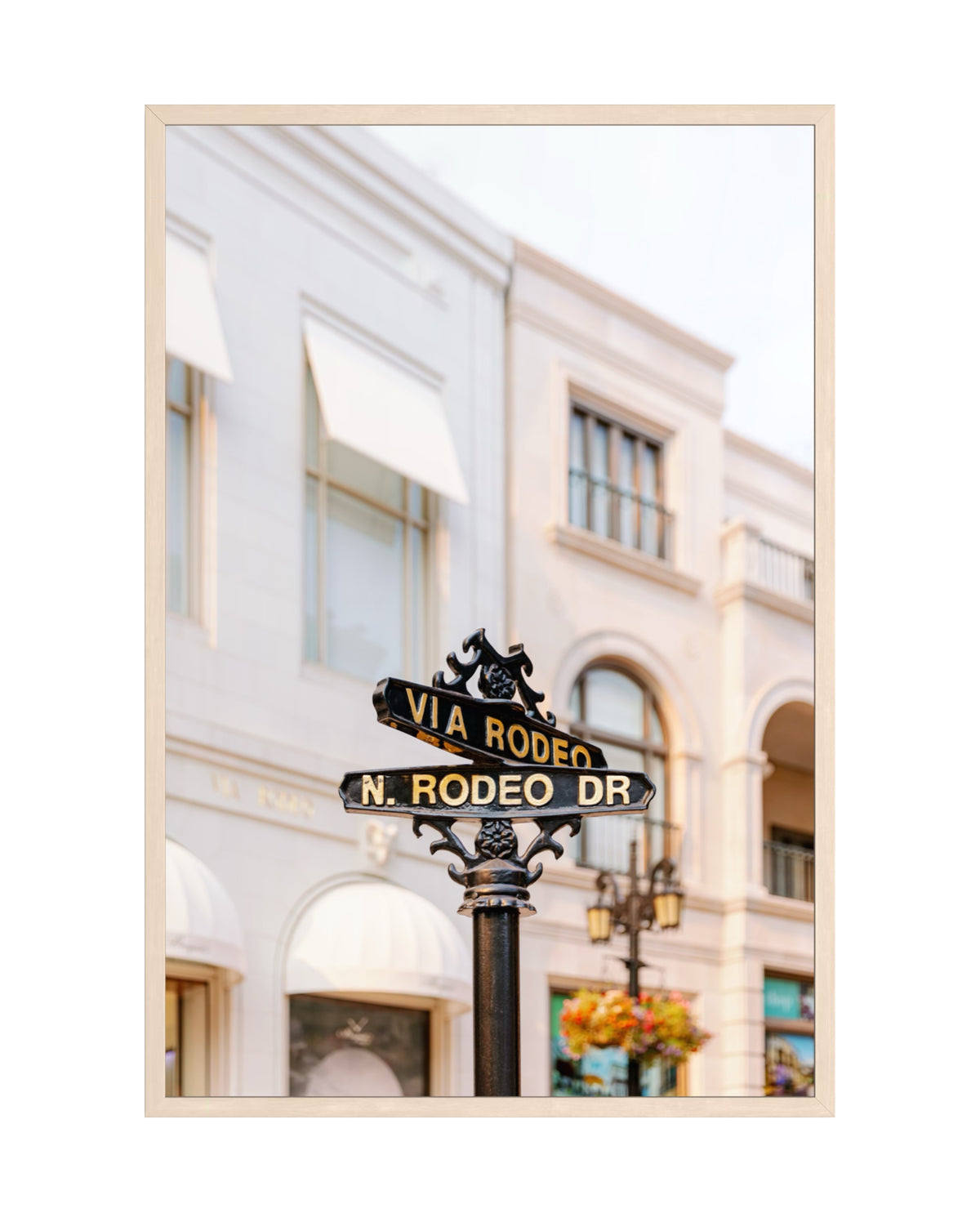 Black Via Rodeo and N. Rodeo Dr street sign in Beverly Hills, set against white buildings on Rodeo Drive.