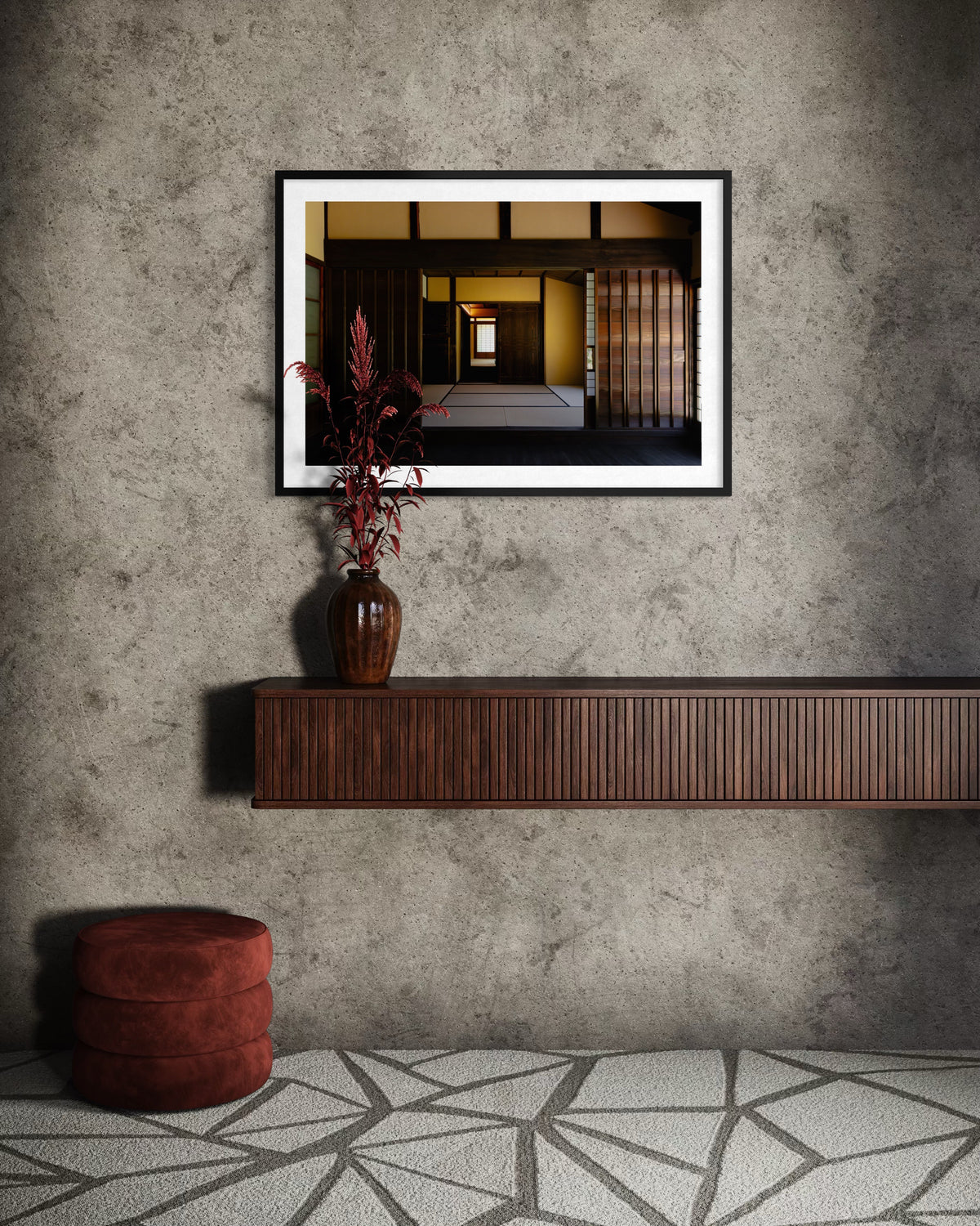 Framed photo above a brown shelf with vase, warm light on red ottoman and gray patterned rug at a Japanese Shōya House.