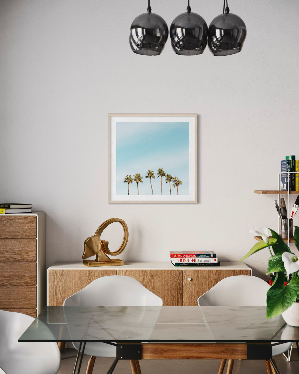Glass dining table and white chairs set beneath hanging lights, with Six Palms artwork on the wall.