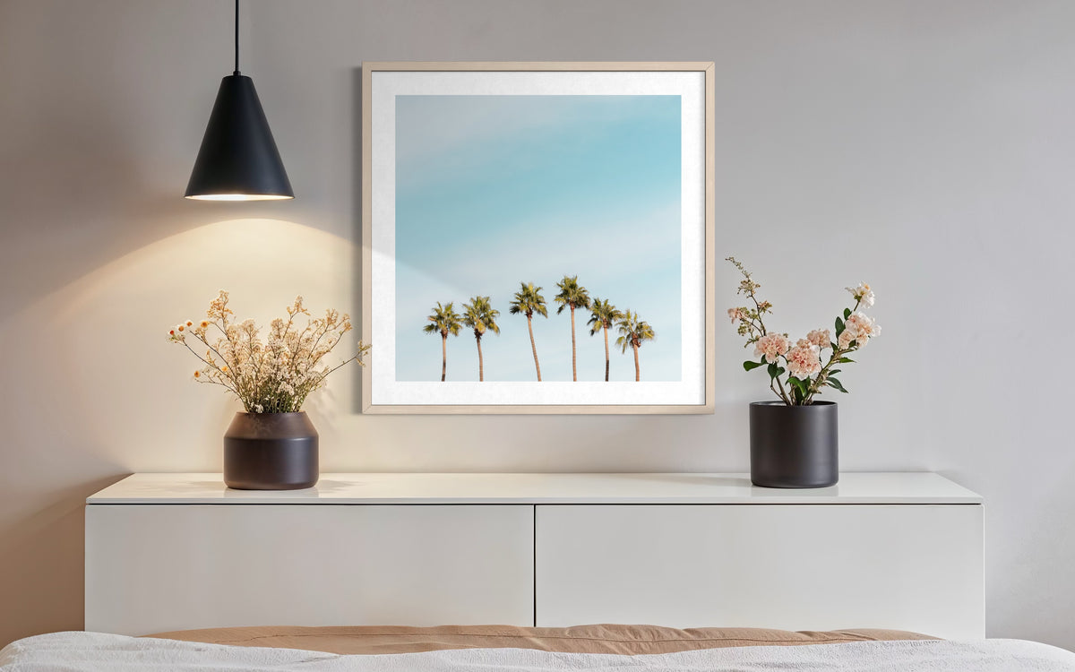 Palm Springs palm trees artwork hangs above a white dresser with vases and a black pendant lamp.