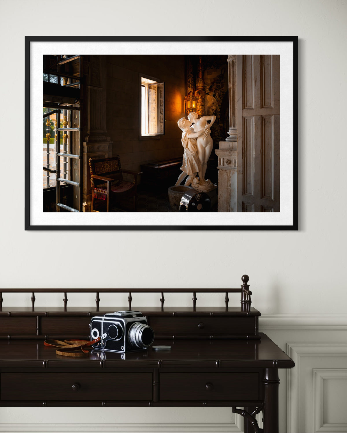 Black-and-white indoor sculpture photo titled “Stolen Kiss” hangs above a wooden desk with a vintage camera.