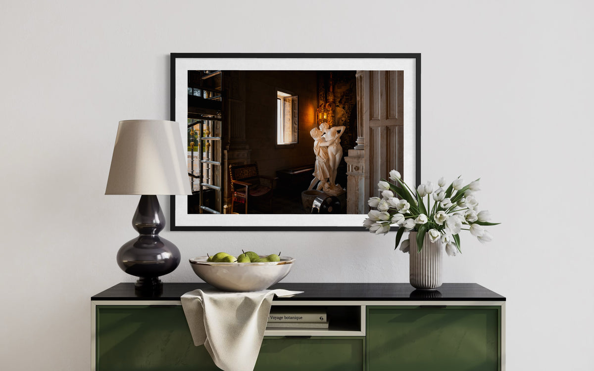 Framed photo of Stolen Kiss sculpture above a black console with a lamp, apples, and white tulips in warm lighting.