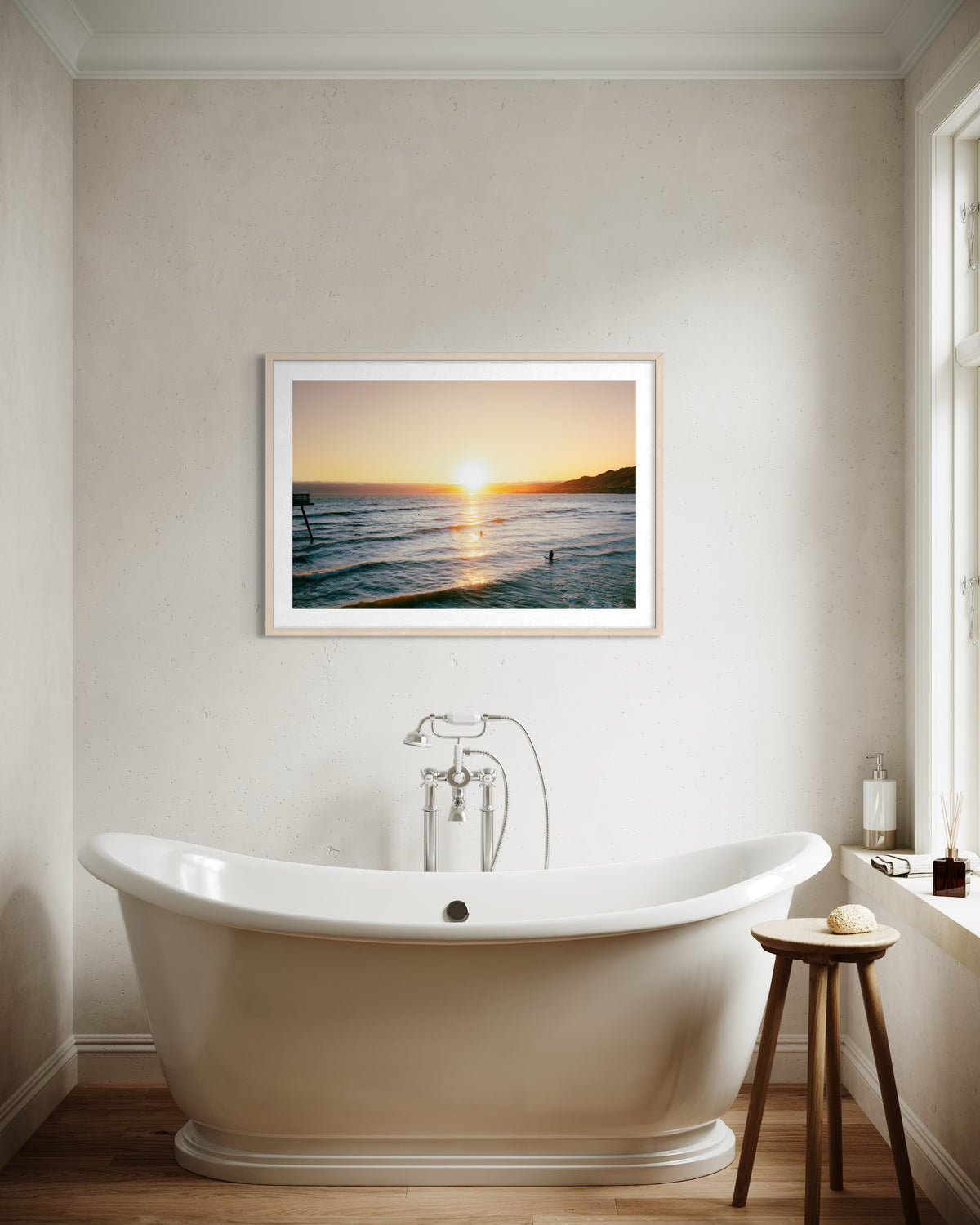 White freestanding tub in bright bathroom with framed “Surfers at Sunset” photograph on the wall.