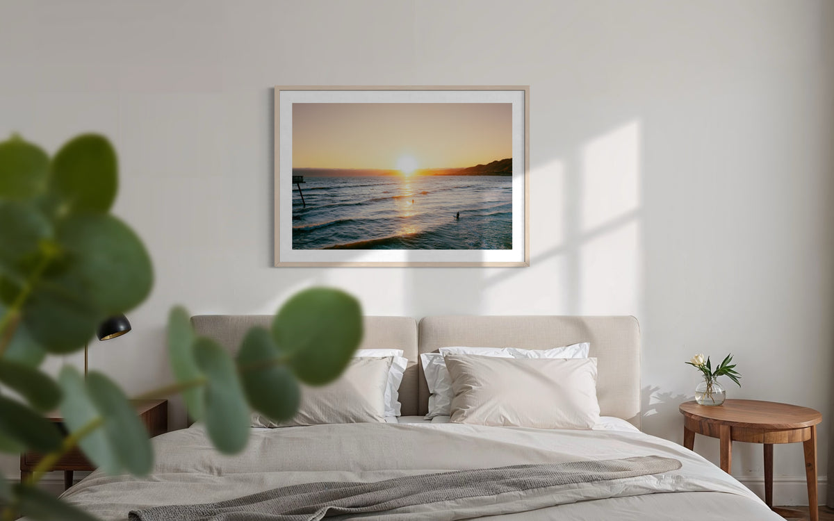Bedroom with “Surfers at Sunset” above the bed, summer evening light coming through a window.