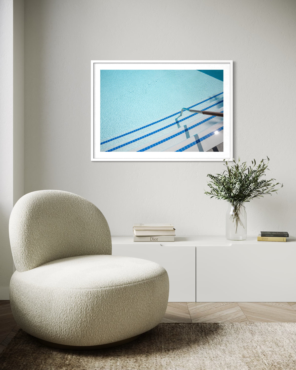 Framed “The Hotel Pool” photo hangs on the wall above a curved white chair, sideboard, and potted plant.
