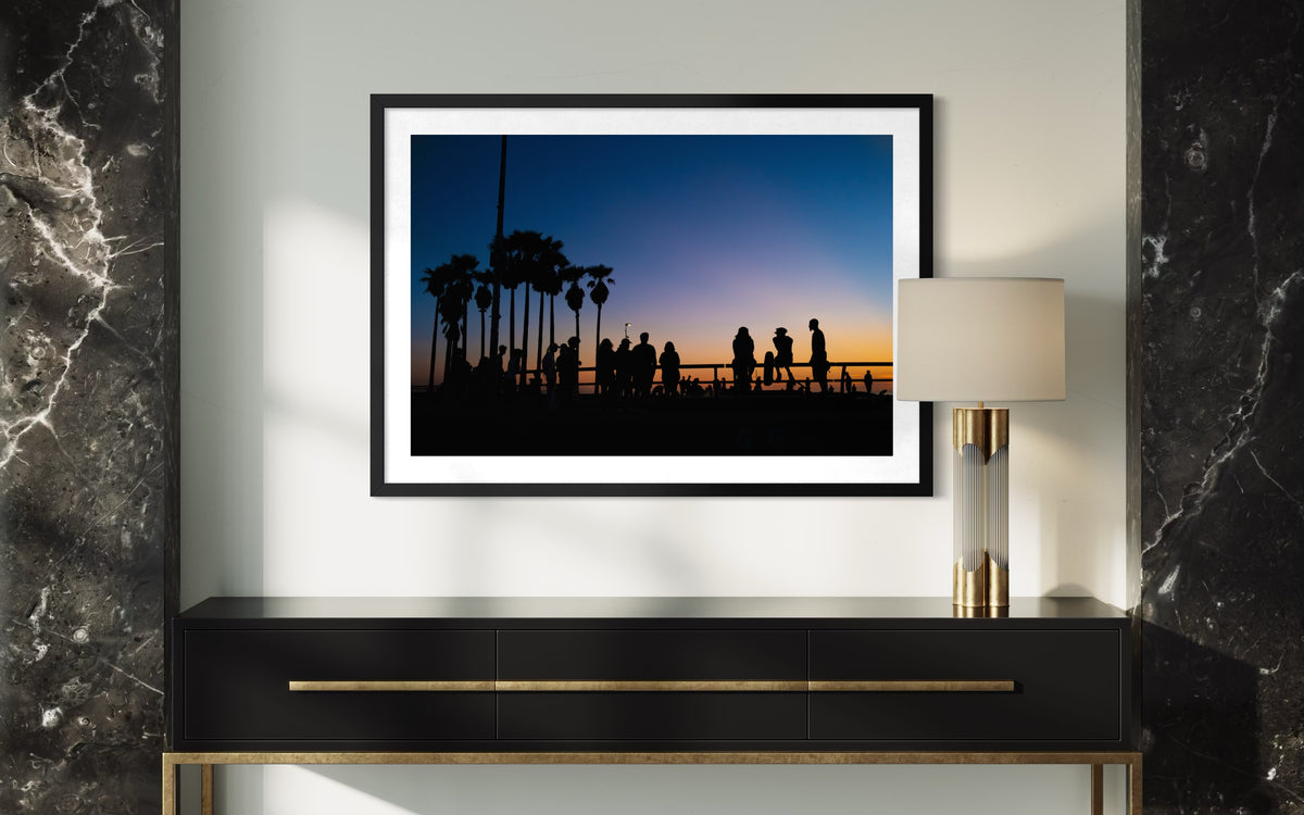 Framed photo of a Venice Beach sunset with palm trees and people, shown above a black console on a marble wall.