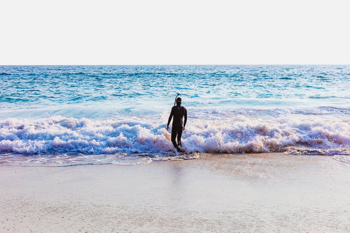 Snorkeler in wetsuit steps from Pacific waves onto sandy shore under a bright sky.