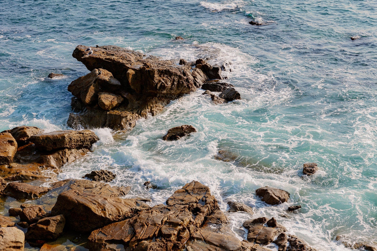 Waves crash against brown rocks and ocean foam glistens along the turquoise Laguna Beach shoreline.