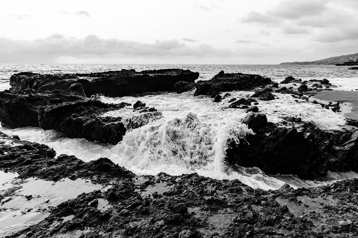 Black-and-white waves crash against rocky coast below a cloudy sky.