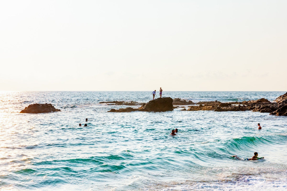 People swimming and relaxing by rocks, with two on a large offshore rock at Laguna Beach in the background.