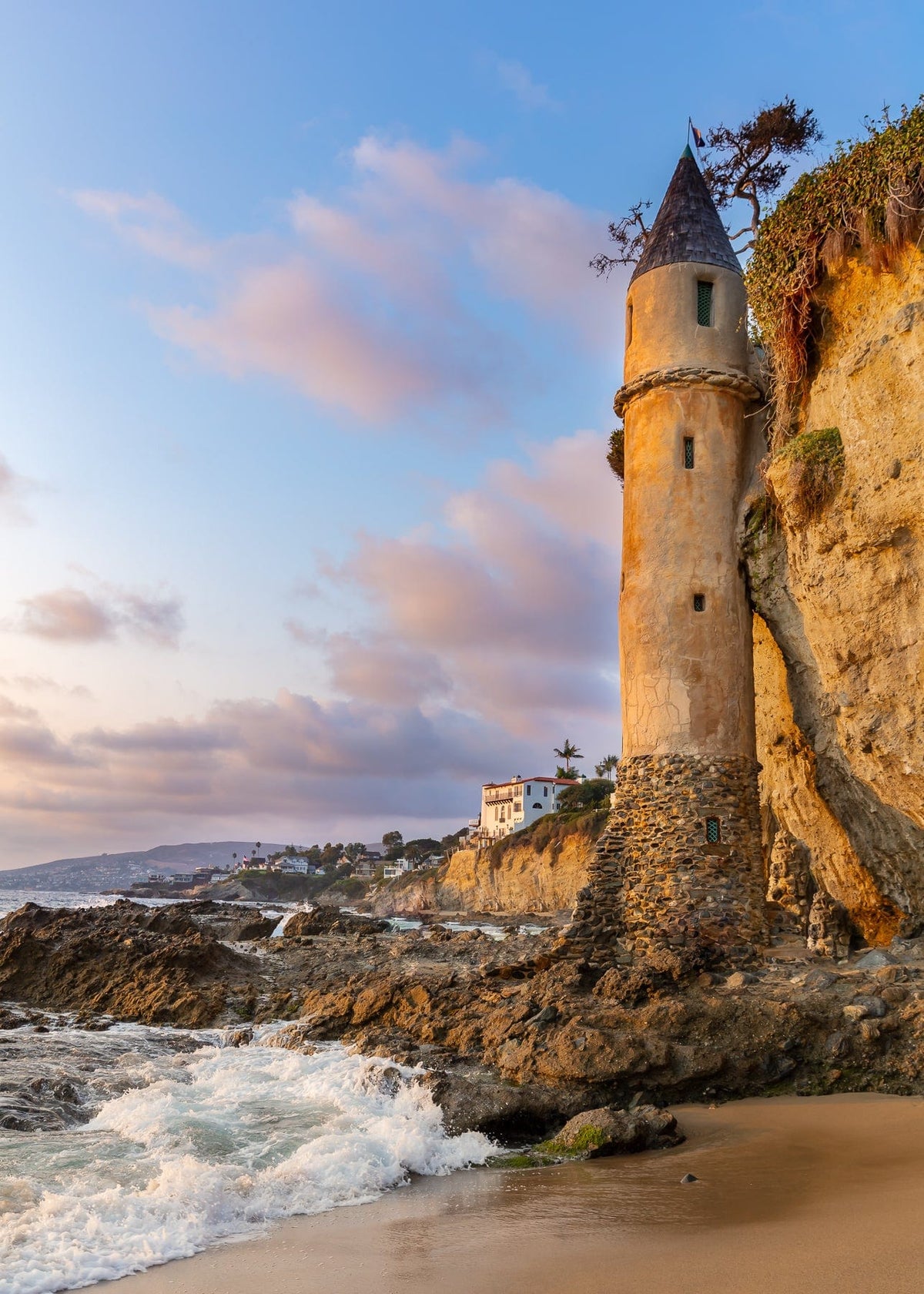 Pirate Tower stands on a rocky Laguna Beach cliff at sunset, with waves crashing below.