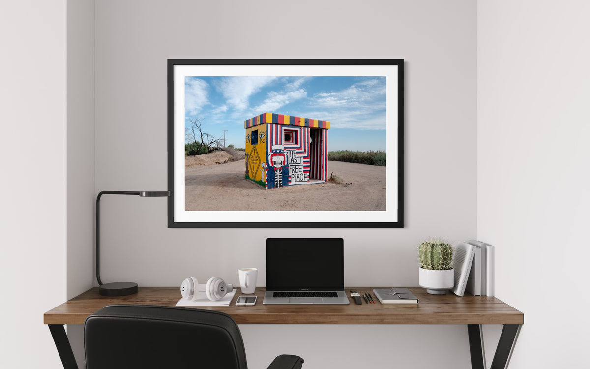 Laptop and plant on desk beneath a framed photo of Slab City’s “The Last Free Place” desert structure.