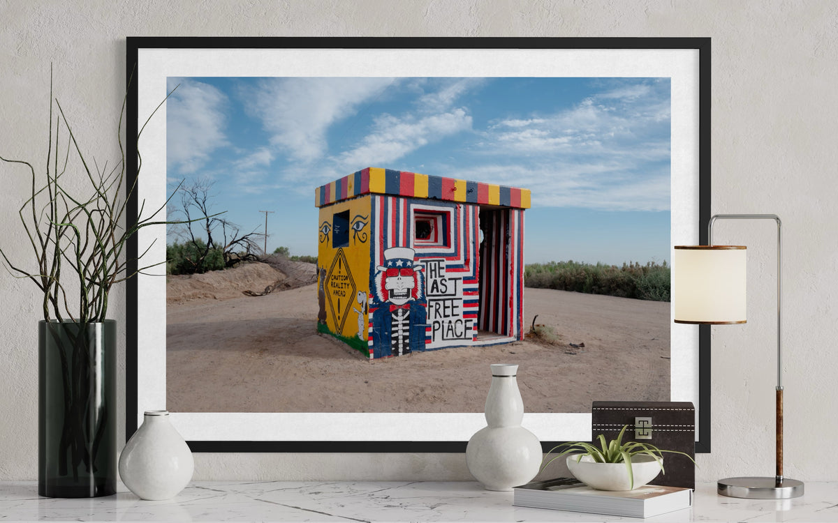 Framed print of “The Last Free Place,” a painted structure in Slab City with table decor in the foreground.