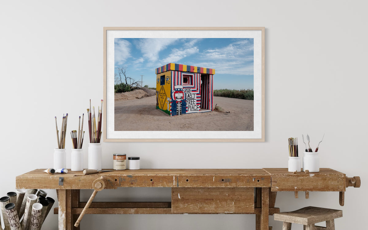 Framed “The Last Free Place” photo on a desert wall above a workbench with art supplies at Slab City.